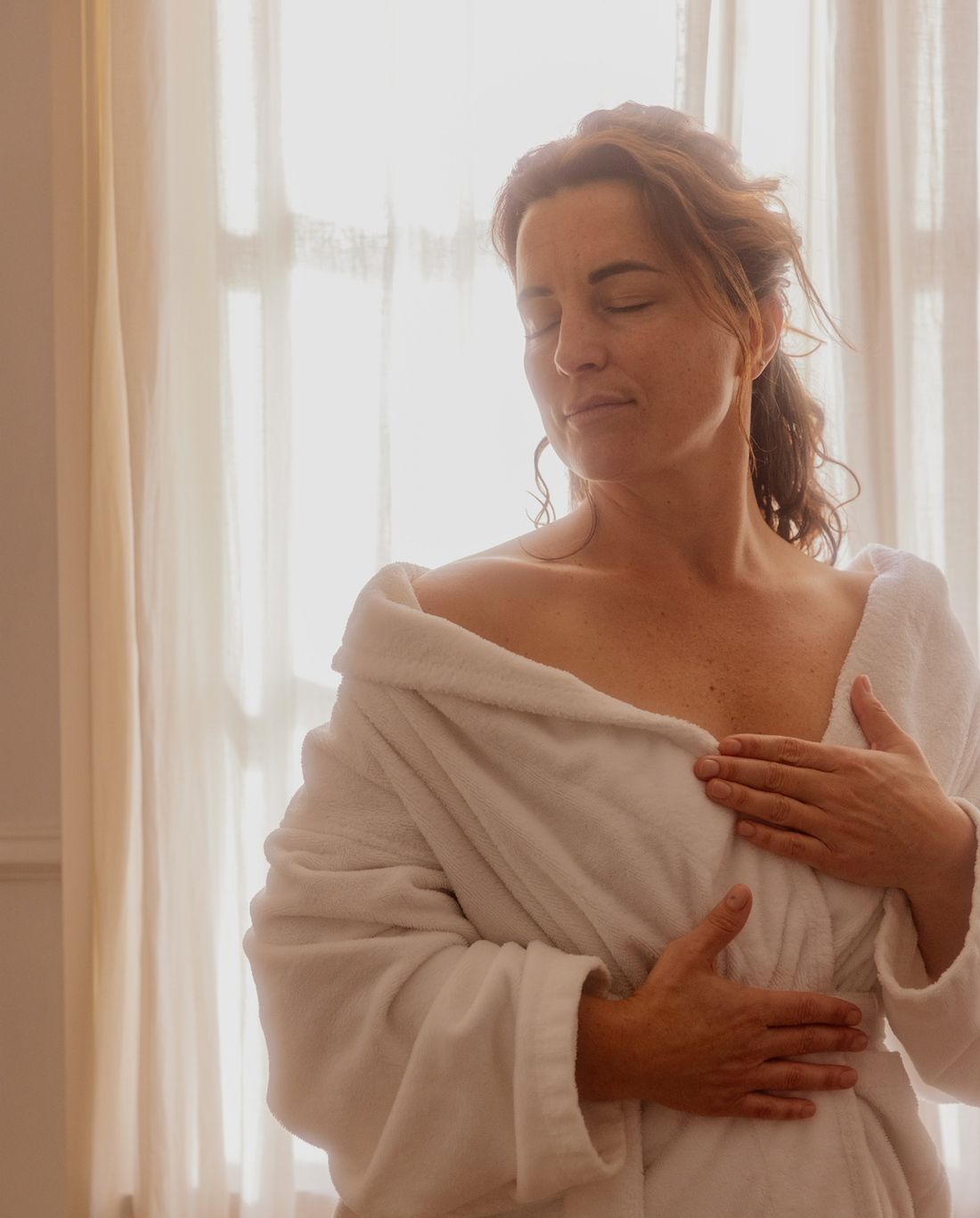 Vrouw in badjas geniet voor een gordijn van de zonnestralen die binnekomen